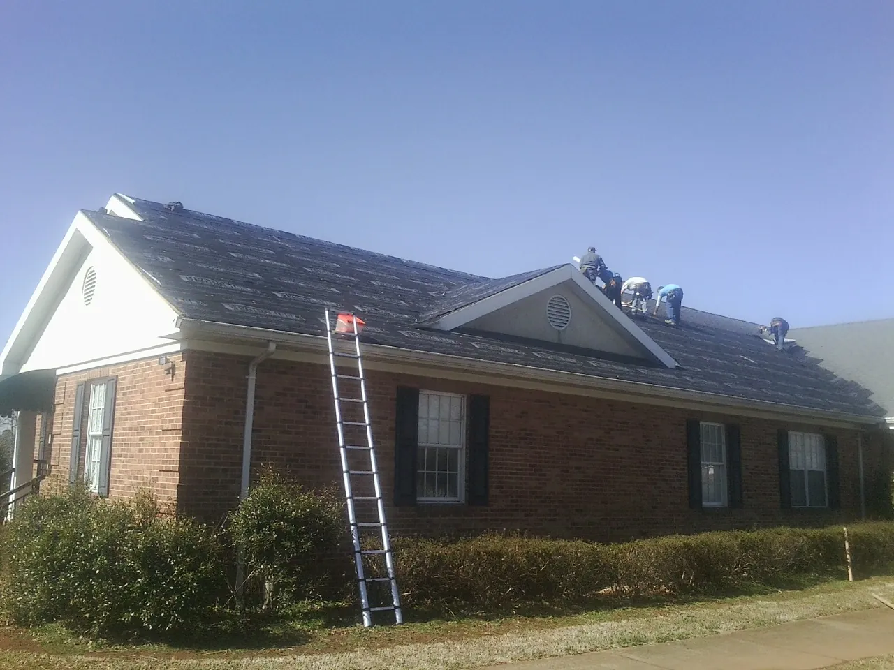 Crew tearing shingles off a brick ranch roof with a ladder set — Metal Roof Repair work by our crew in Rutland